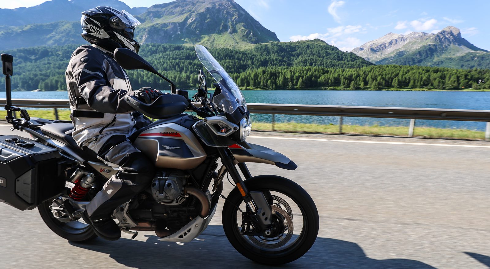 Motard équipé d'un casque intégral et d'une veste moto roulant sur une route de montagne au bord d'un lac